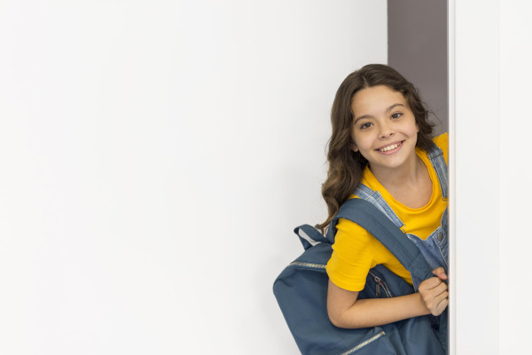A smiling child with a backpack peeks around a doorway